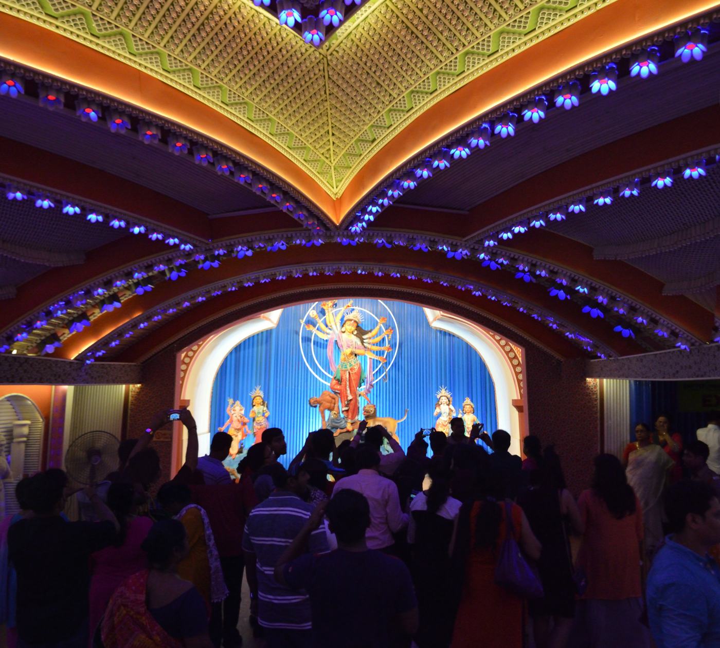 Inside a lit pandal at night, the goddess on a blue stage, people gathered before her